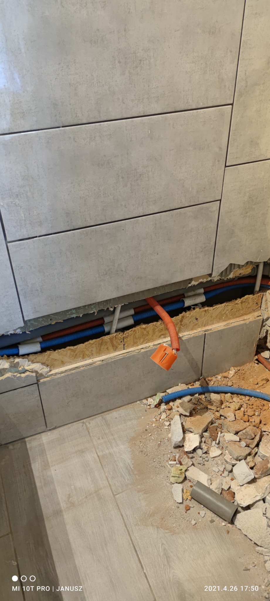 Fragment ściany z odsłoniętymi instalacjami hydraulicznymi i elektrycznymi w trakcie remontu, widoczne rury w kolorach niebieskim, pomarańczowym i białym, gruz i pył na podłodze.