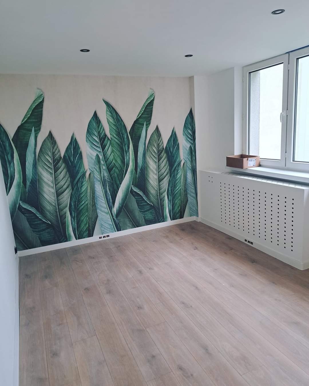 Pokój po remoncie z fototapetą z motywem tropikalnych liści, jasną drewnianą podłogą i białym grzejnikiem pod oknem.