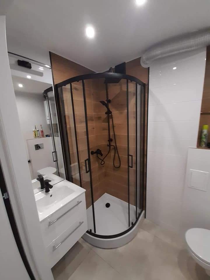 Nowoczesna łazienka z narożną kabiną prysznicową z czarnymi profilami, białą umywalką z szafką i płytkami imitującymi drewno na ścianie wewnątrz kabiny.