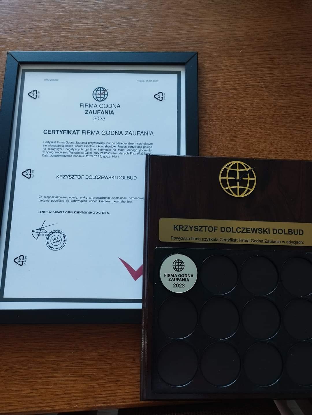 Certyfikat 'Firma Godna Zaufania 2023' w czarnej ramce, obok drewniana tabliczka z logo firmy i pustymi polami na kolejne certyfikaty, na drewnianym tle.