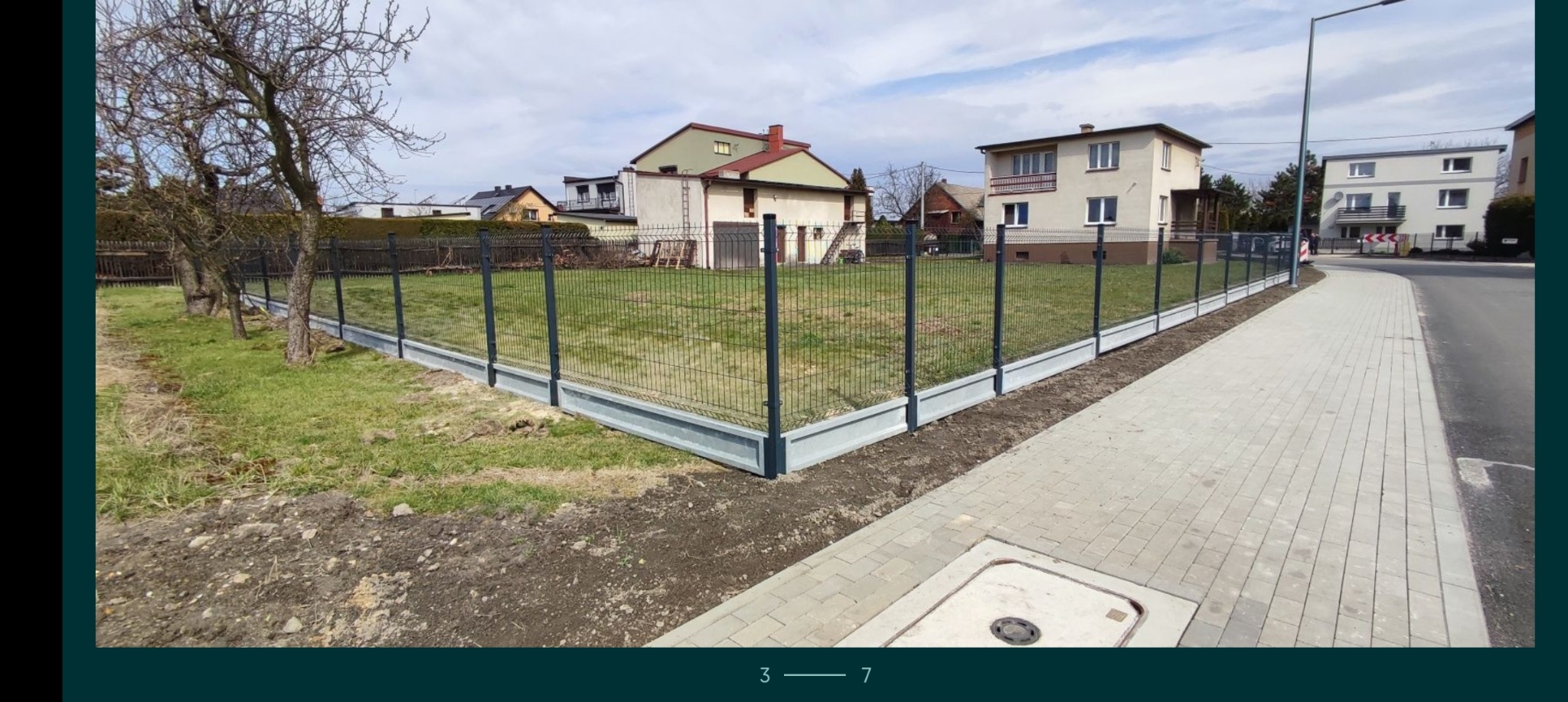 Nowoczesne ogrodzenie panelowe z podmurówką betonową, widok z zewnątrz na tle domów i drzew bez liści, przylegające do chodnika z kostki.