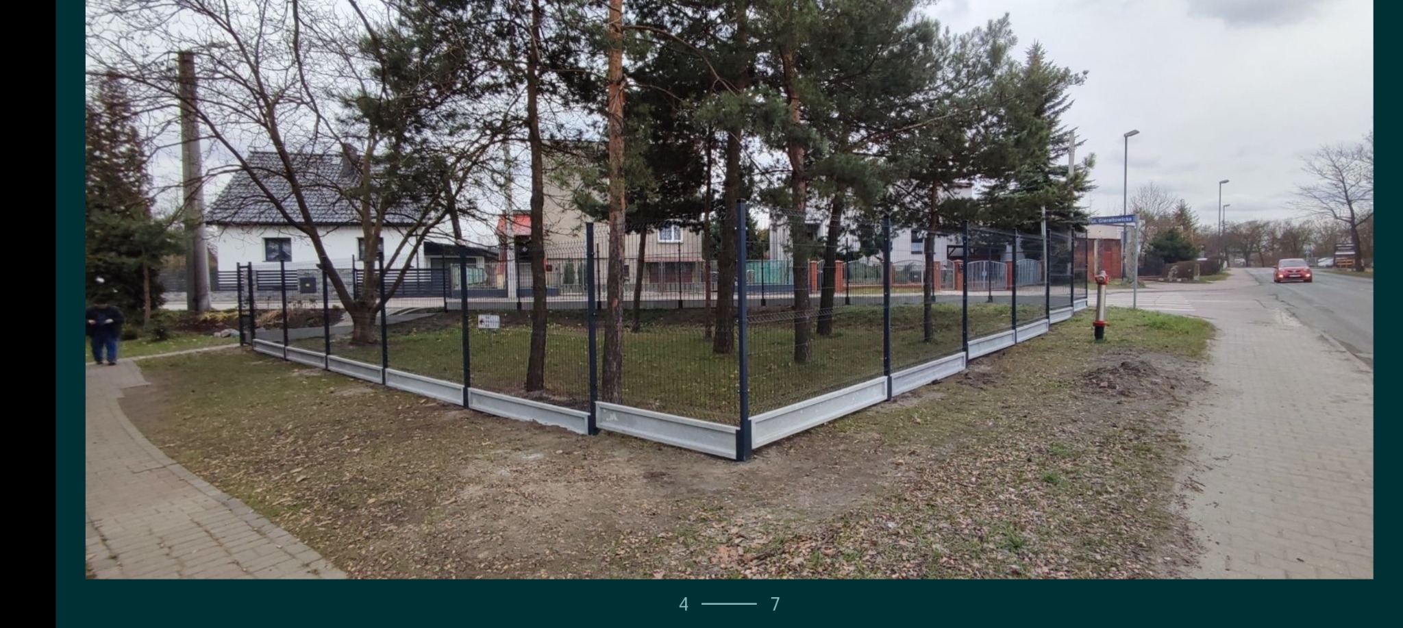 Nowoczesne ogrodzenie panelowe z podmurówką betonową, otoczone drzewami iglastymi, widok z chodnika w pochmurny dzień.