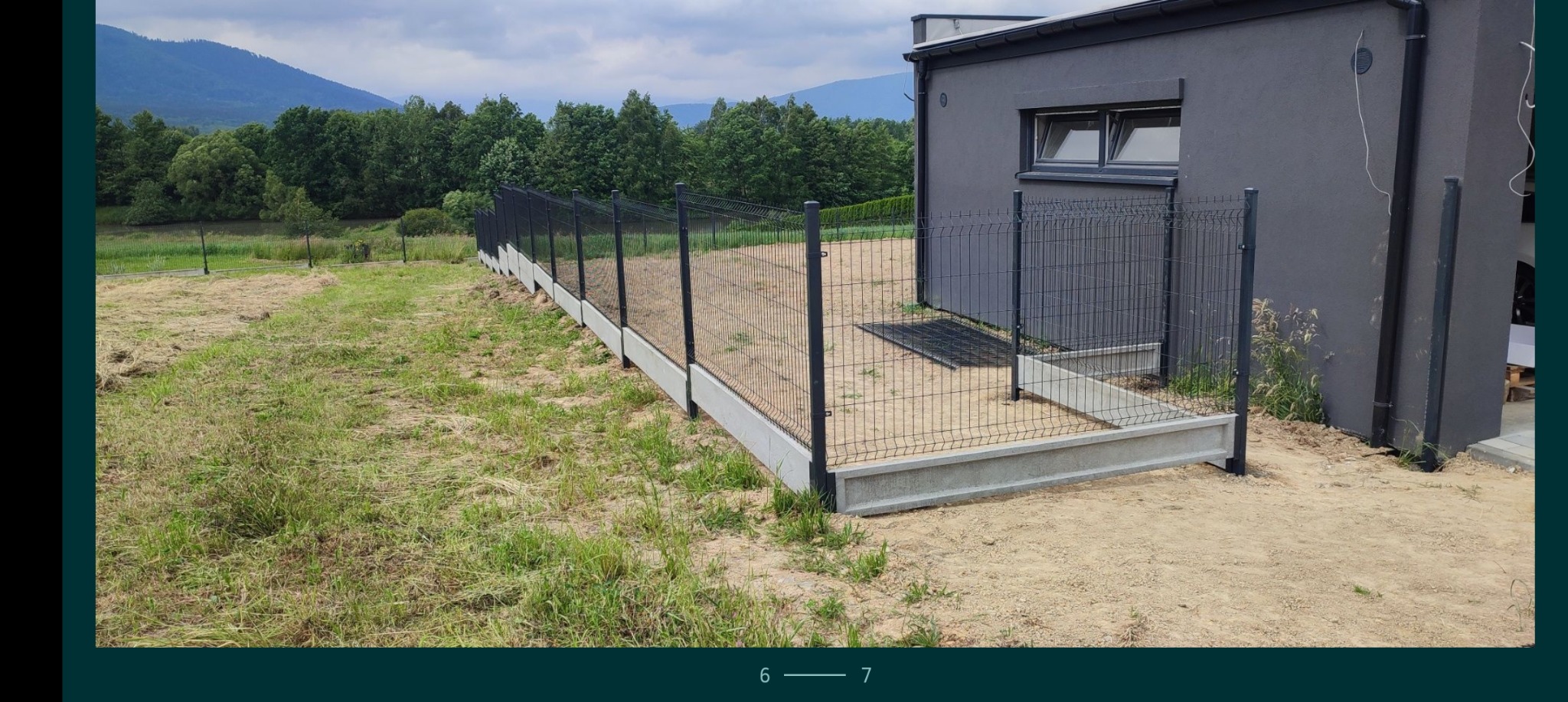 Czarny metalowy płot panelowy z betonową podmurówką na nierównym terenie, widok na szary dom i zalesione wzgórza w tle.