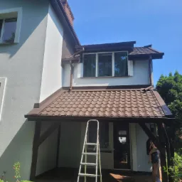 Dwupiętrowy dom z jasną elewacją i brązowym dachem, widoczna aluminiowa drabina oparta o ścianę, kobieta z dzieckiem na tarasie.