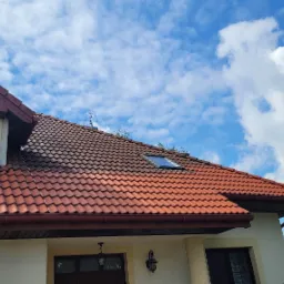 Częściowo wyczyszczony dach z czerwonej dachówki, widoczna wyraźna różnica w kolorze pomiędzy czystą a brudną powierzchnią, na tle błękitnego nieba z chmurami.