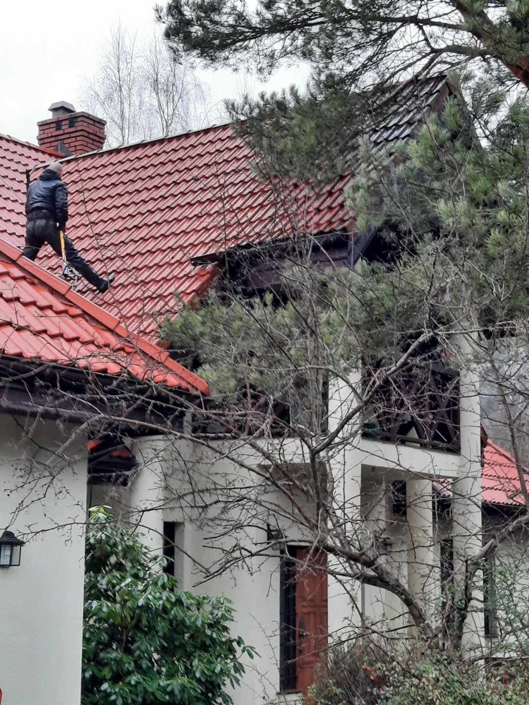 Pracownik w ciemnej kurtce i spodniach na czerwonym dachu domu z ceglanym kominem, trzymający żółty młotek, widok z dołu przez gałęzie drzew.