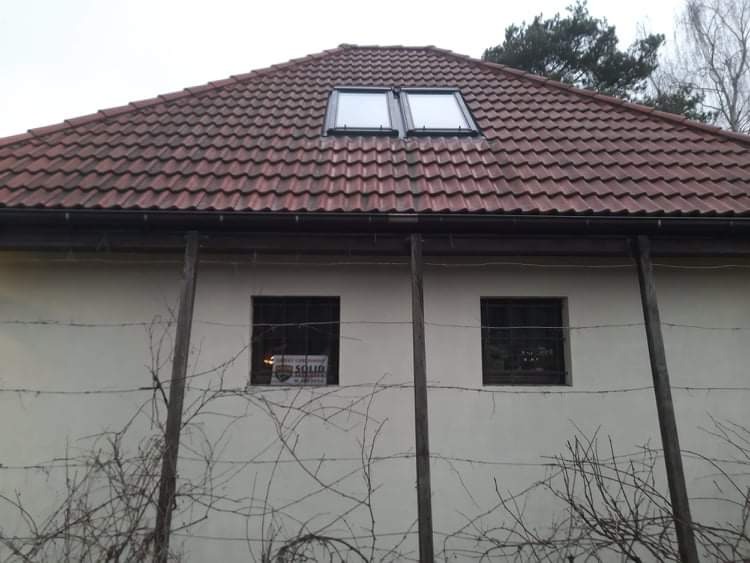 Dom z czerwonym dachem, dwoma oknami dachowymi i jasną elewacją, z widocznymi rynnami i oknami zabezpieczonymi kratami, częściowo zasłonięty przez suchą roślinność i drut kolczasty.