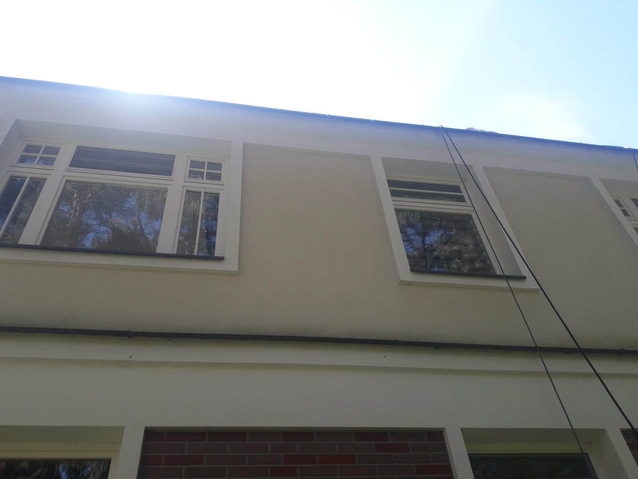 Jasna elewacja budynku z widocznymi oknami i elementami dekoracyjnymi, sfotografowana pod kątem z dołu, z linami zwisającymi z dachu.