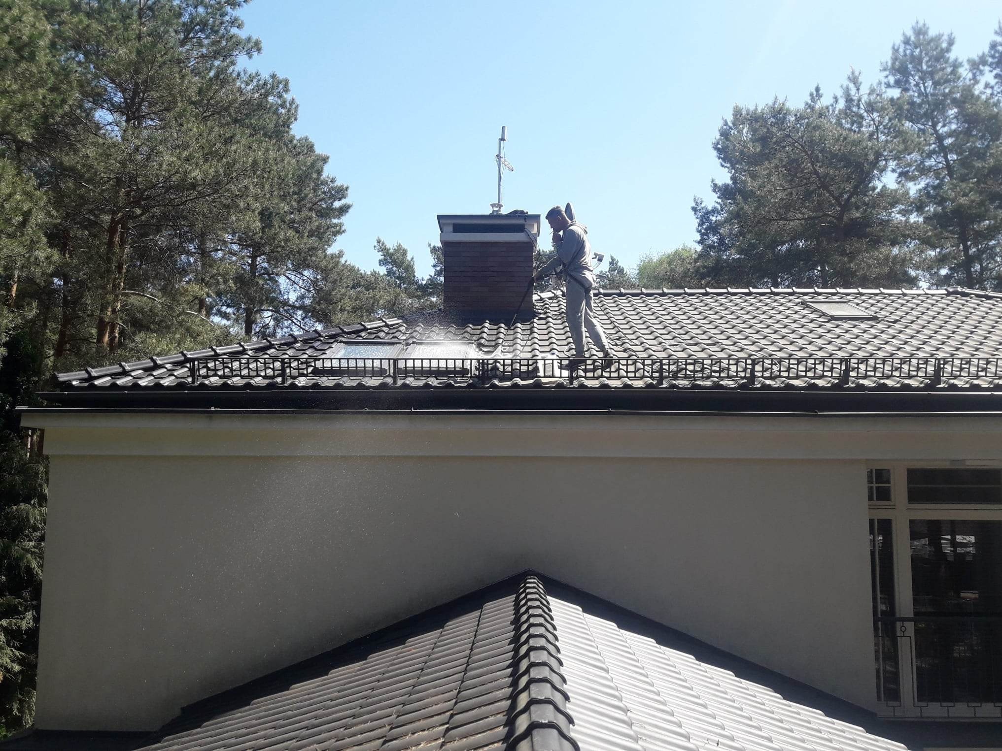 Pracownik w kombinezonie ochronnym spryskuje dach z dachówki ceramicznej w pobliżu komina z cegły, widoczne drzewa w tle.