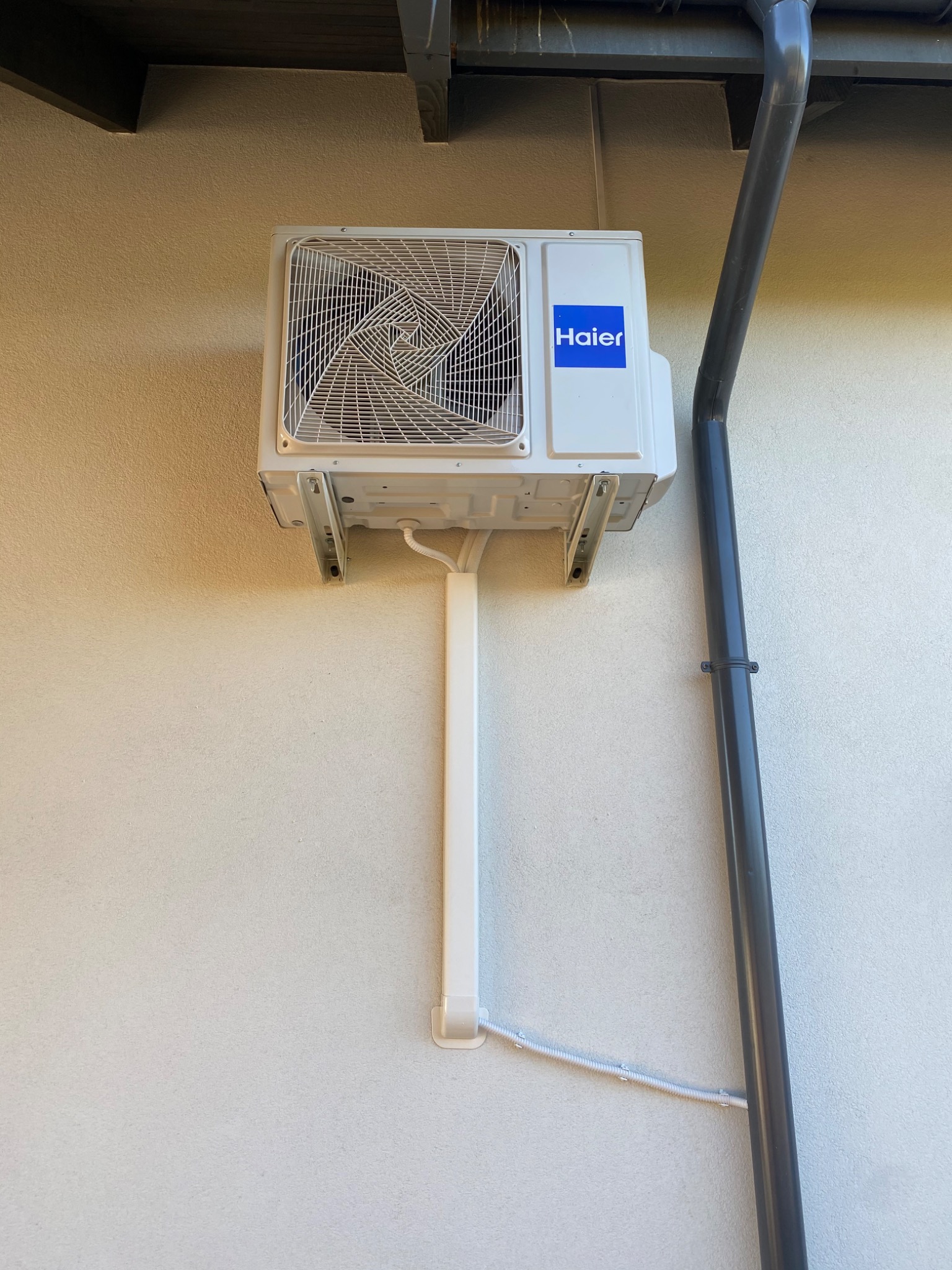 Biała jednostka zewnętrzna klimatyzatora Haier zamontowana na jasnej, teksturowanej ścianie budynku, z widoczną rurą odprowadzającą skropliny i szarą rynną po prawej stronie.