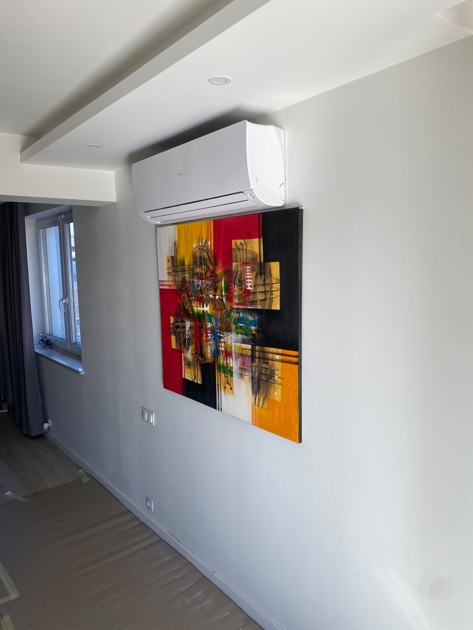 Biały klimatyzator Haier zamontowany na ścianie nad abstrakcyjnym obrazem w salonie z oknem i zasłoną; podłoga zabezpieczona folią malarską podczas montażu.