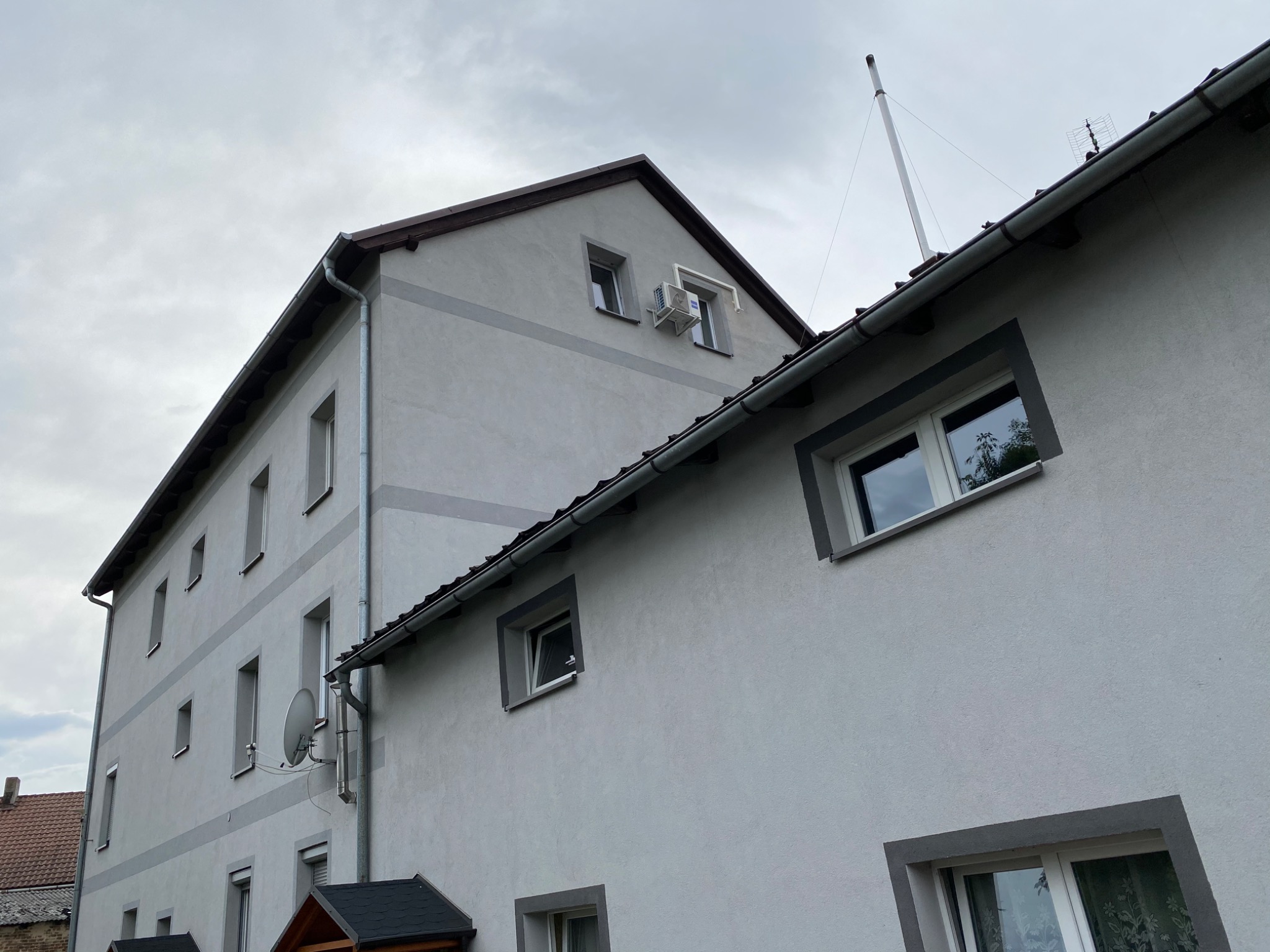 Szary budynek wielorodzinny z zamontowaną klimatyzacją zewnętrzną pod oknem na poddaszu, widoczna antena satelitarna i komin na dachu.