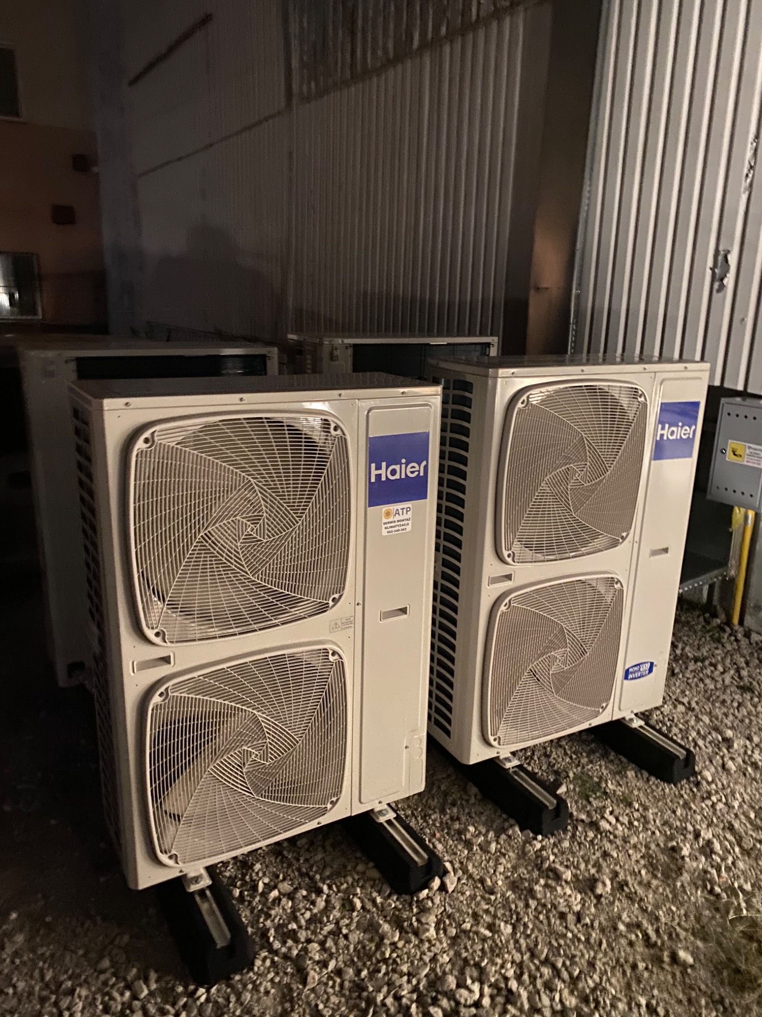 Dwie jednostki zewnętrzne klimatyzacji marki Haier ustawione na żwirowym podłożu, widoczne logo i naklejki informacyjne, tło stanowi blaszana ściana.