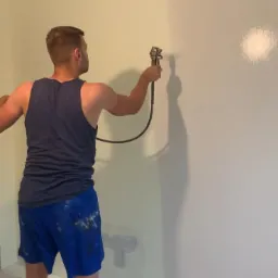 Mężczyzna w trakcie natryskowego nakładania farby na ścianę w pomieszczeniu, widoczne ślady farby na spodenkach.