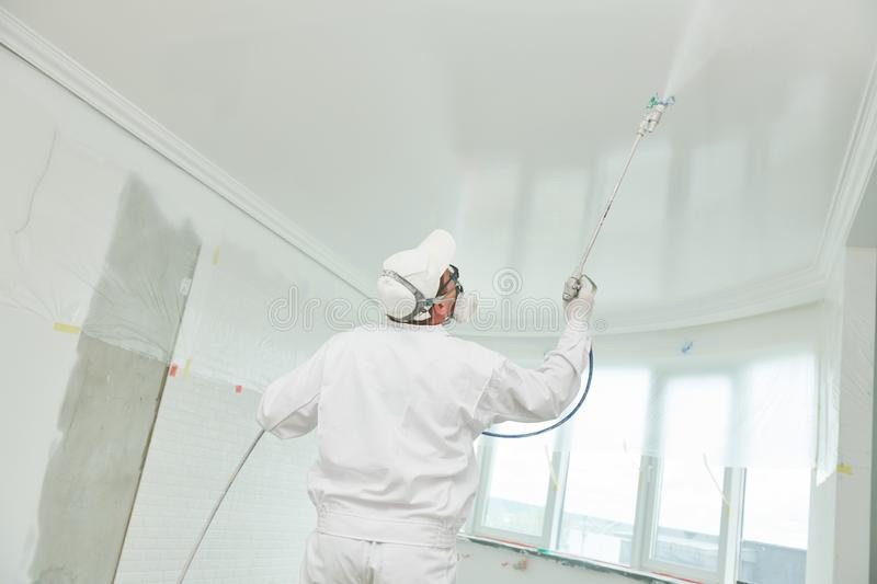 Pracownik w kombinezonie ochronnym, z maską i rękawicami, natryskowo maluje sufit w jasnym pomieszczeniu z oknami, widoczne zabezpieczenia folią na ścianach.