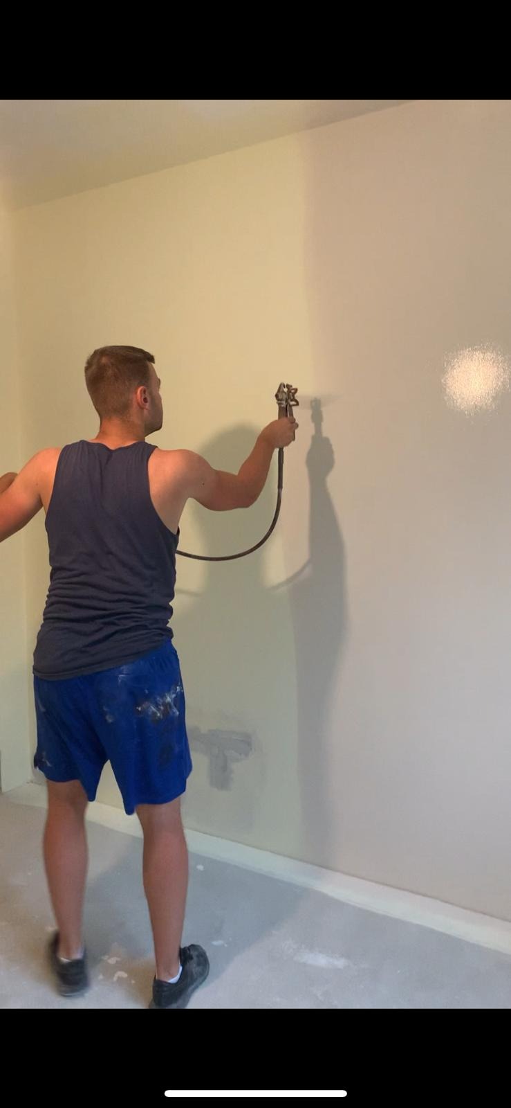 Mężczyzna w trakcie natryskowego nakładania farby na ścianę w pomieszczeniu, widoczne ślady farby na spodenkach.