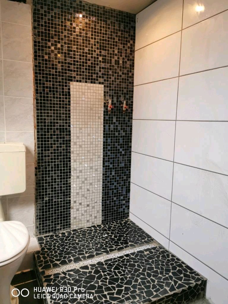 Wykończona kabina prysznicowa z mozaiką czarno-białą na ścianie i podłodze, widoczny odpływ liniowy, białe płytki na sąsiedniej ścianie i fragment toalety.