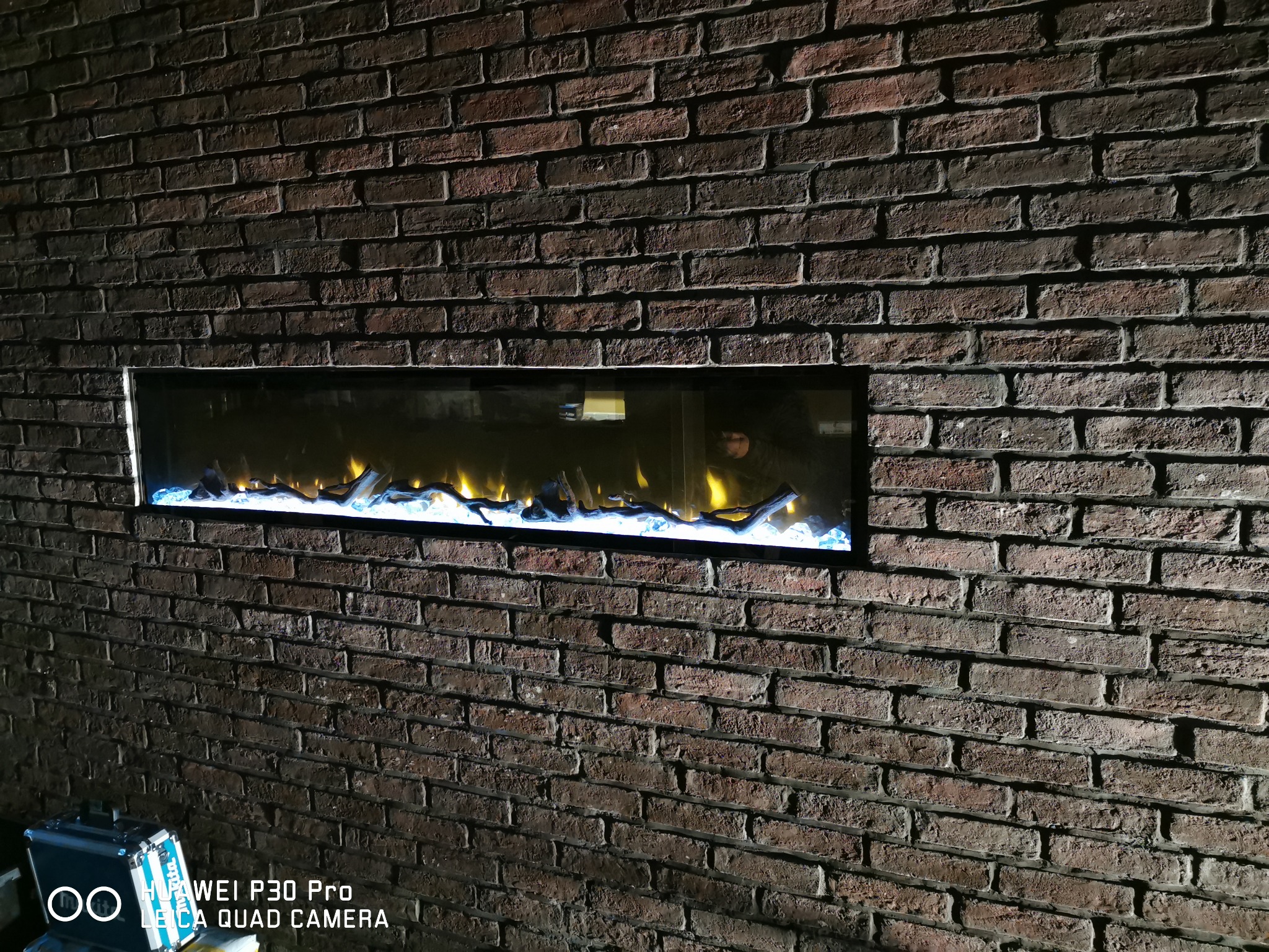 Wnęka w ścianie z cegły z zainstalowanym elektrycznym kominkiem z efektem płomieni i dekoracyjnymi elementami imitującymi drewno i kryształki lodu.