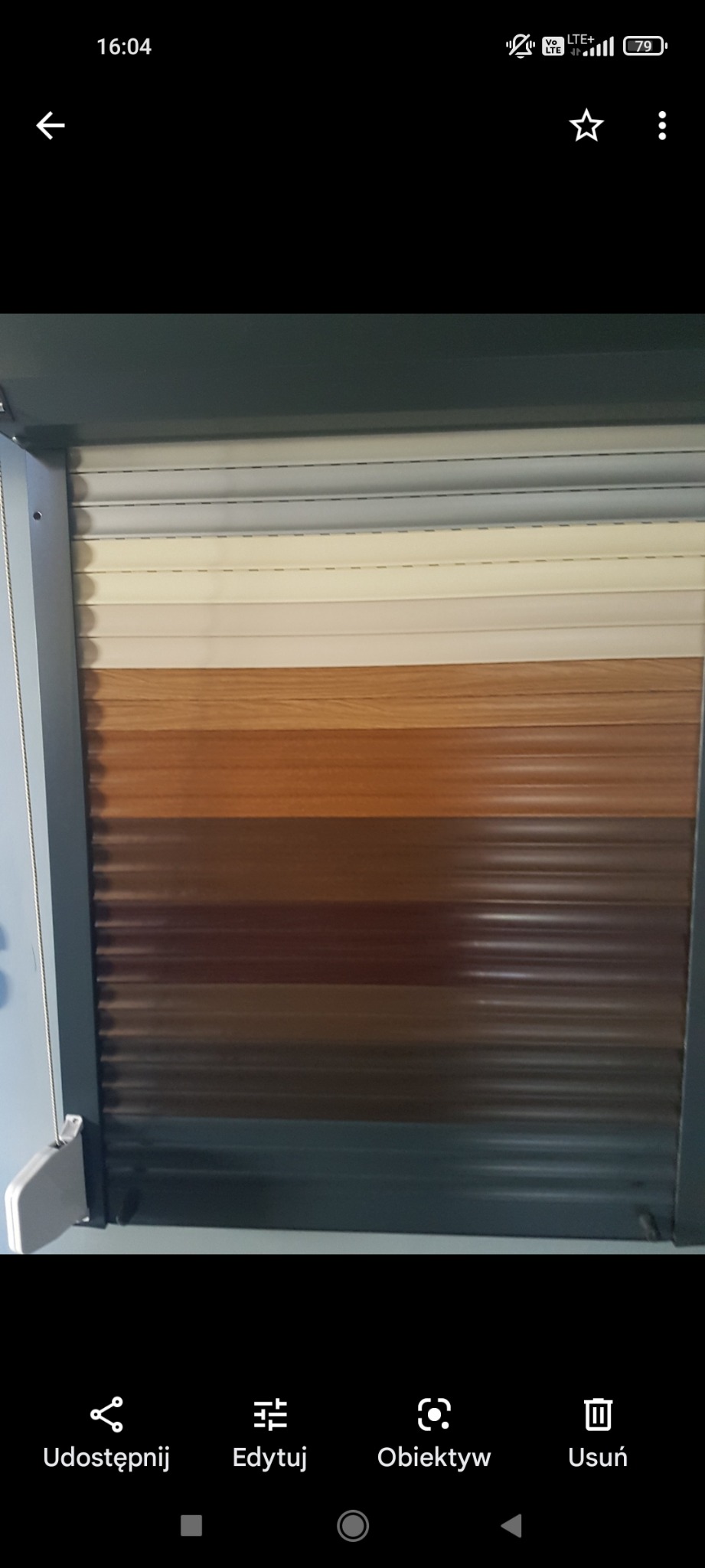 Fragment zamkniętej rolety okiennej, prezentującej różne odcienie brązu i beżu, z widocznym mechanizmem zwijania po lewej stronie.