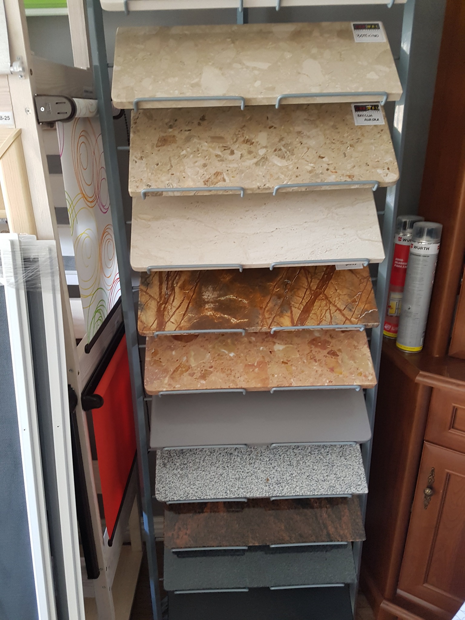 Ekspozycja próbek różnych kolorów i faktur materiałów wykończeniowych, umieszczonych na metalowym stojaku w sklepie, obok widoczne butelki z preparatami Wurth i fragment ramy okiennej