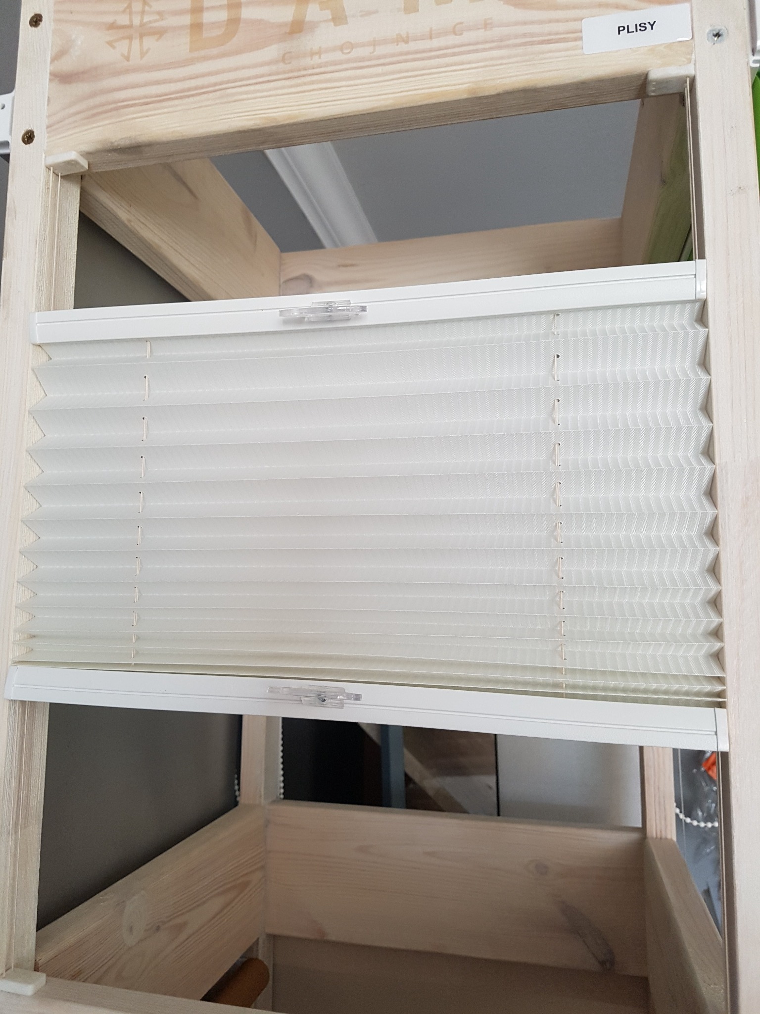 Zamontowana, biała, plisowana roleta okienna w drewnianej ramie, widoczna etykieta z napisem 'Plisy'.