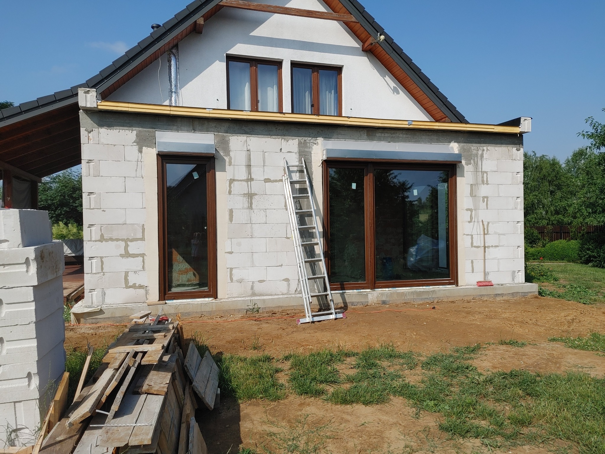 Budynek w trakcie budowy z zamontowanymi oknami z brązowymi ramami, widoczna drabina oparta o ścianę, stos bloczków i desek na pierwszym planie.
