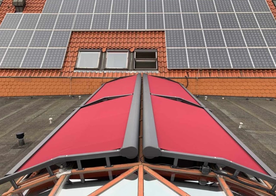 Czerwona, składana markiza dachowa z szarymi elementami konstrukcyjnymi, widoczna z dołu, z panelami słonecznymi i oknami na dachu w tle.