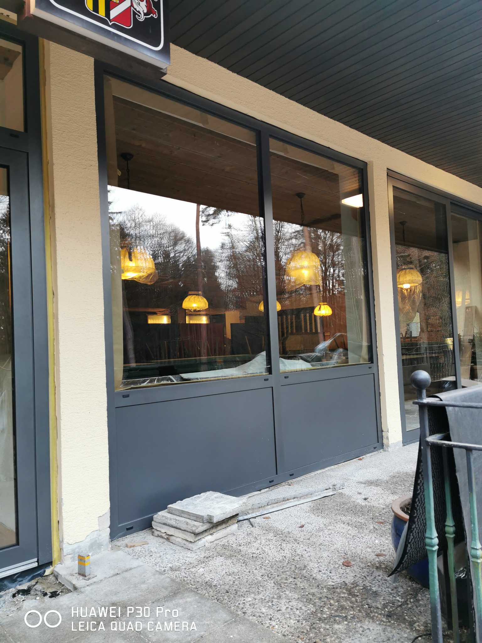 Duże, przeszklone okno z szarymi ramami i dolną częścią, widok na wnętrze z wiszącymi lampami, stos płyt chodnikowych leżący na zewnątrz, element elewacji w kolorze beżowym.