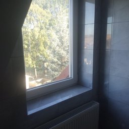 Remont Dla Ciebie - Wykończona ściana w łazience z szarymi płytkami ceramicznymi, białe okno z widokiem na drzewa, grzejnik pod oknem.
