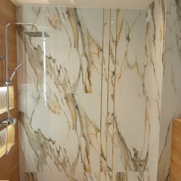 Remont Dla Ciebie - Nowoczesna kabina prysznicowa z marmurowymi płytkami i złotymi akcentami, drewnopodobne wykończenie podłogi i zabudowy. Oświetlenie sufitowe.