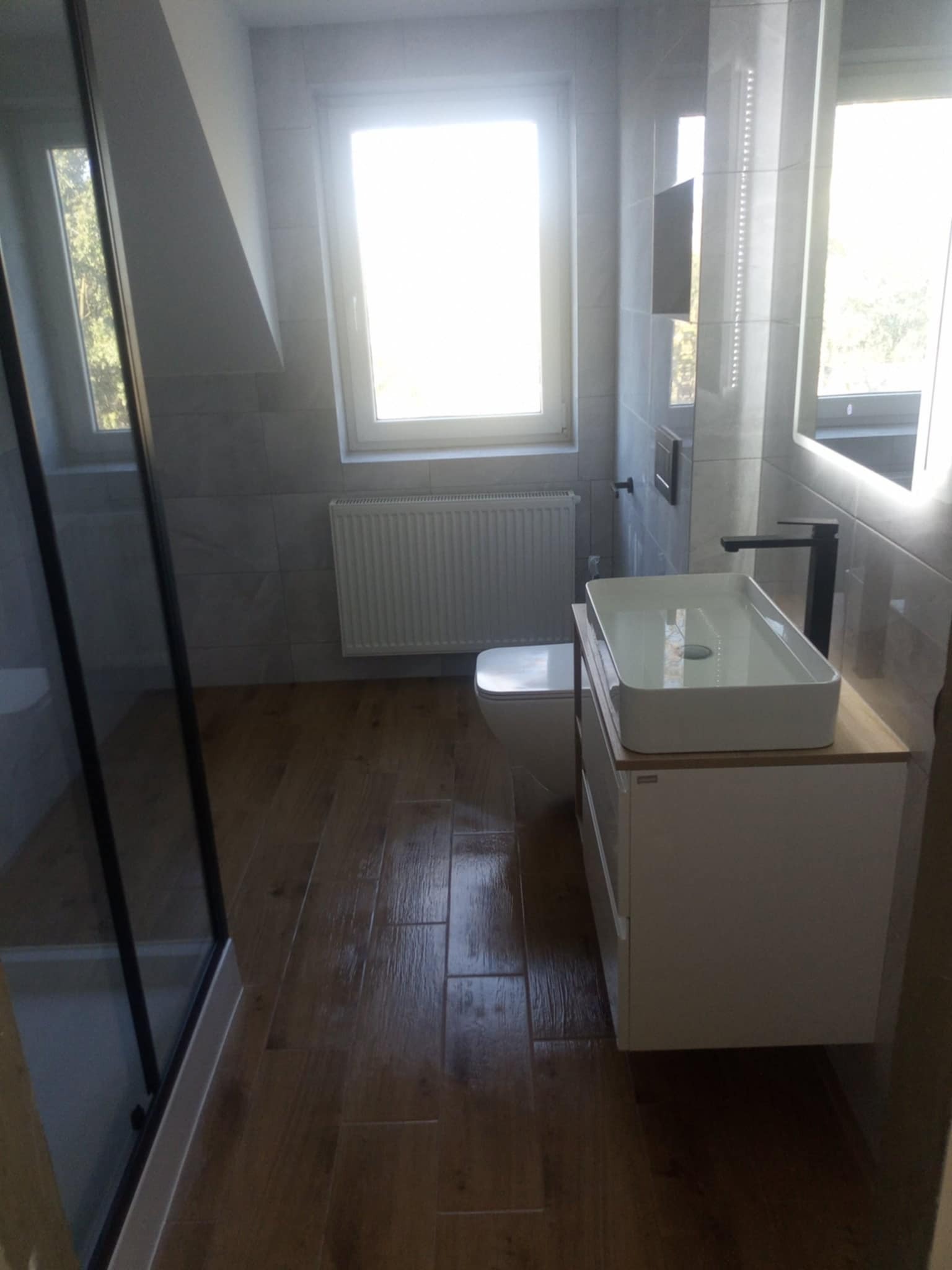 Nowoczesna łazienka z drewnopodobną podłogą, prostokątną umywalką nablatową z czarną armaturą, oświetlonym lustrem i grzejnikiem panelowym pod oknem.