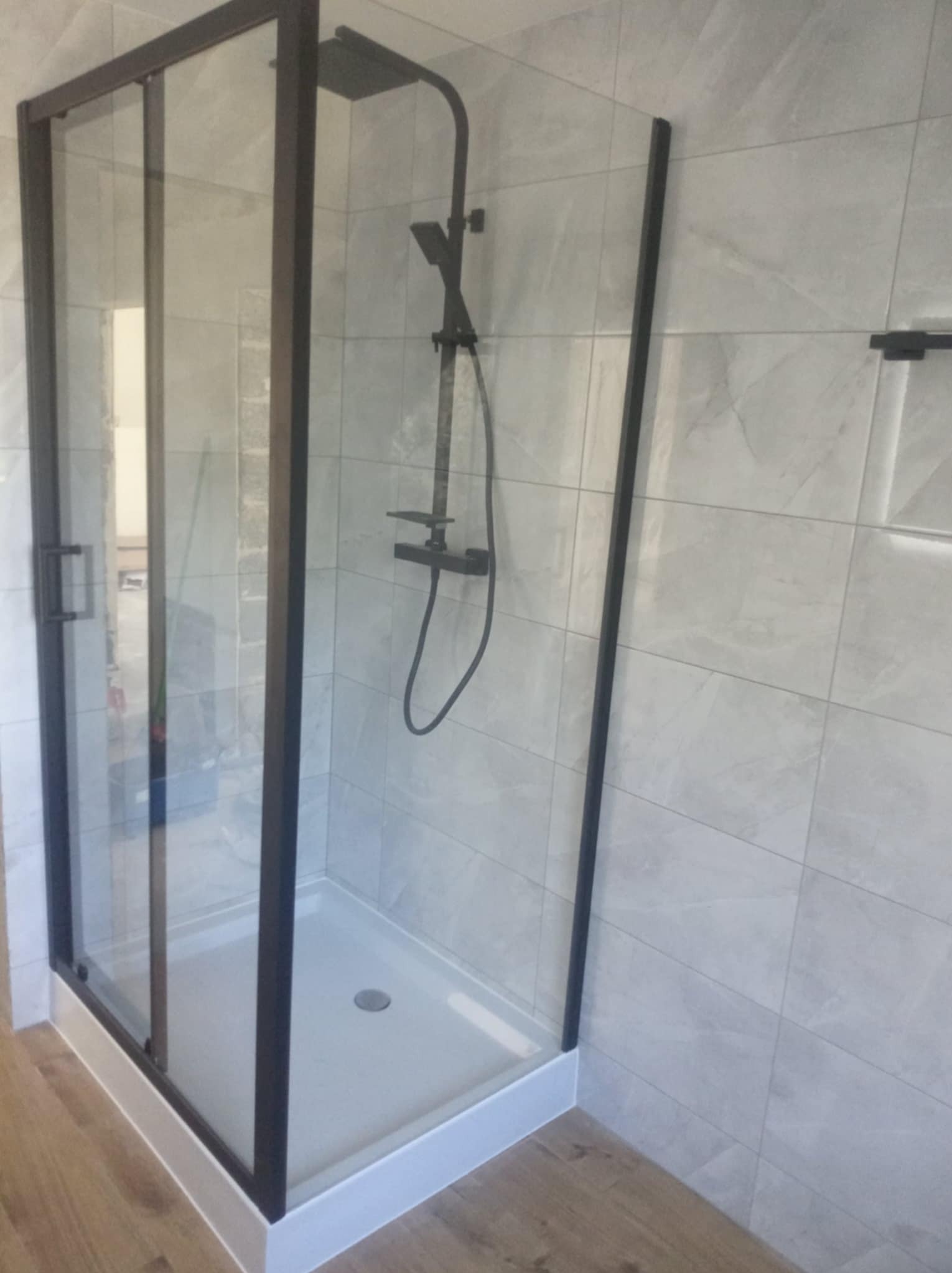 Nowoczesna kabina prysznicowa z czarnymi profilami i deszczownicą, jasne płytki imitujące marmur na ścianach, drewnopodobna podłoga.