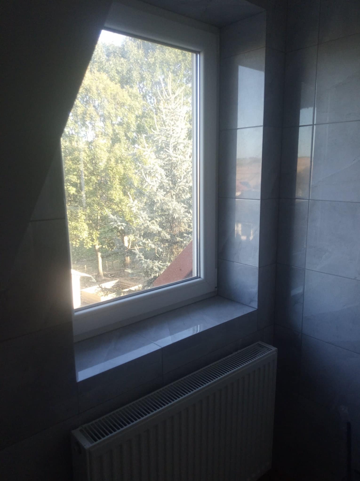 Wykończona ściana w łazience z szarymi płytkami ceramicznymi, białe okno z widokiem na drzewa, grzejnik pod oknem.