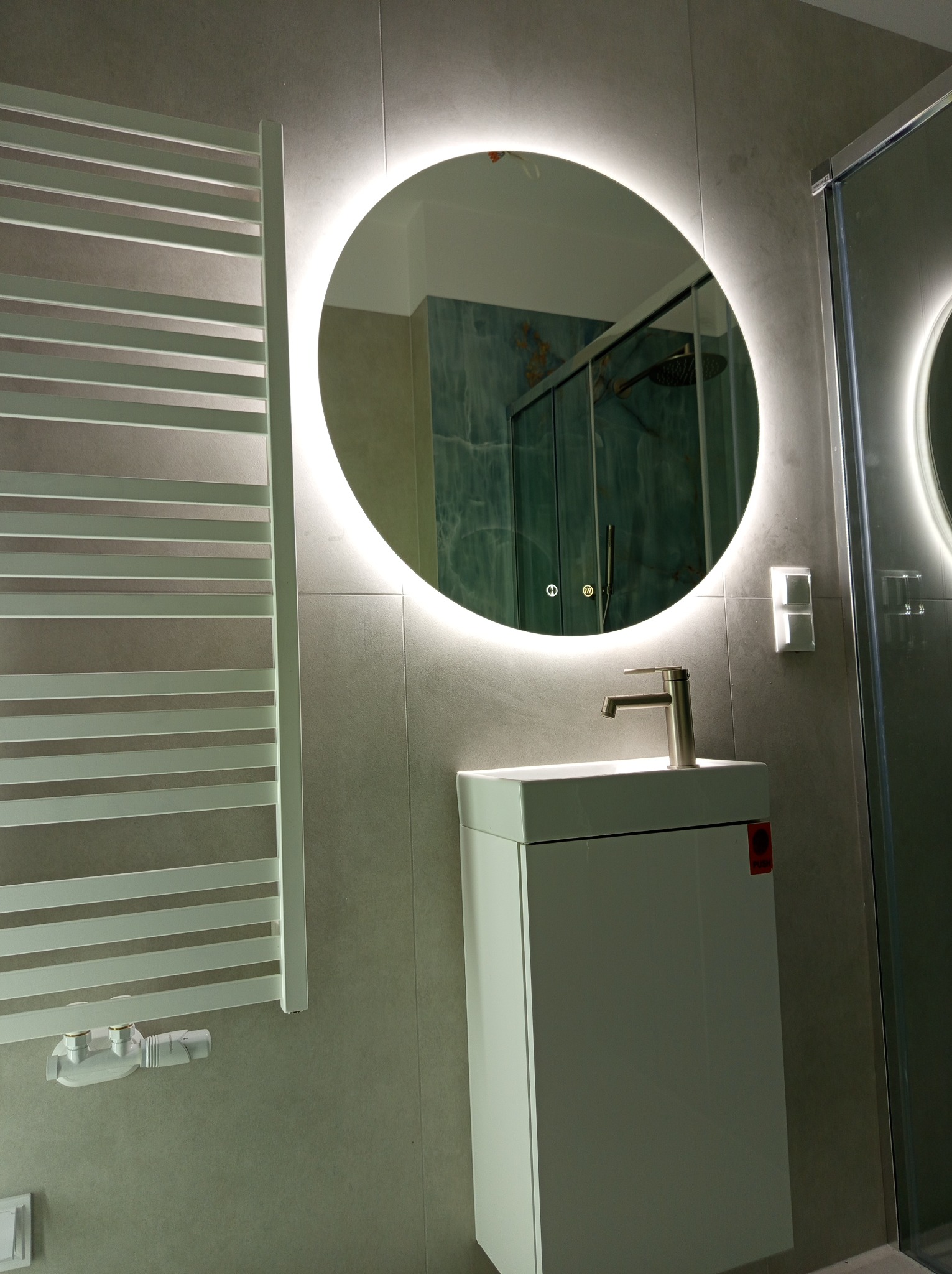 Nowoczesna łazienka z okrągłym lustrem LED, białą szafką pod umywalką i grzejnikiem drabinkowym. Szare płytki ścienne i kabina prysznicowa w tle.