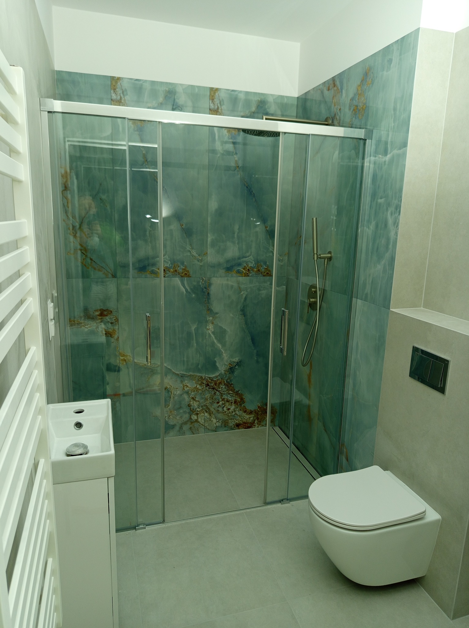 Nowoczesna łazienka z kabiną prysznicową z przesuwnymi drzwiami, umywalką na szafce i podwieszaną toaletą. Ściany wyłożone płytkami imitującymi marmur.