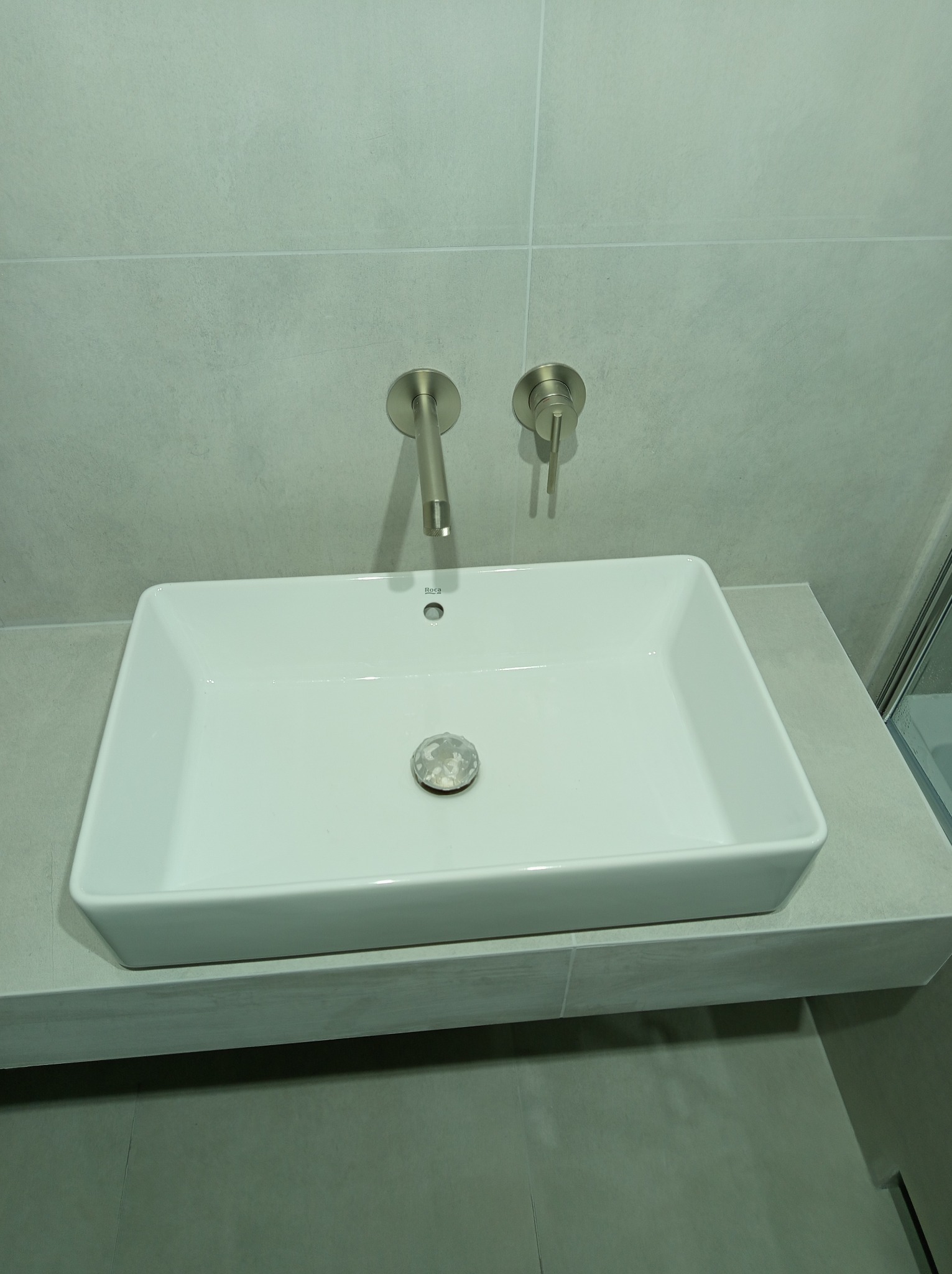 Biała, prostokątna umywalka Roca na blacie z płytek, z oryginalnymi, srebrnymi bateriami ściennymi na tle szarych płytek. Nowoczesna łazienka.