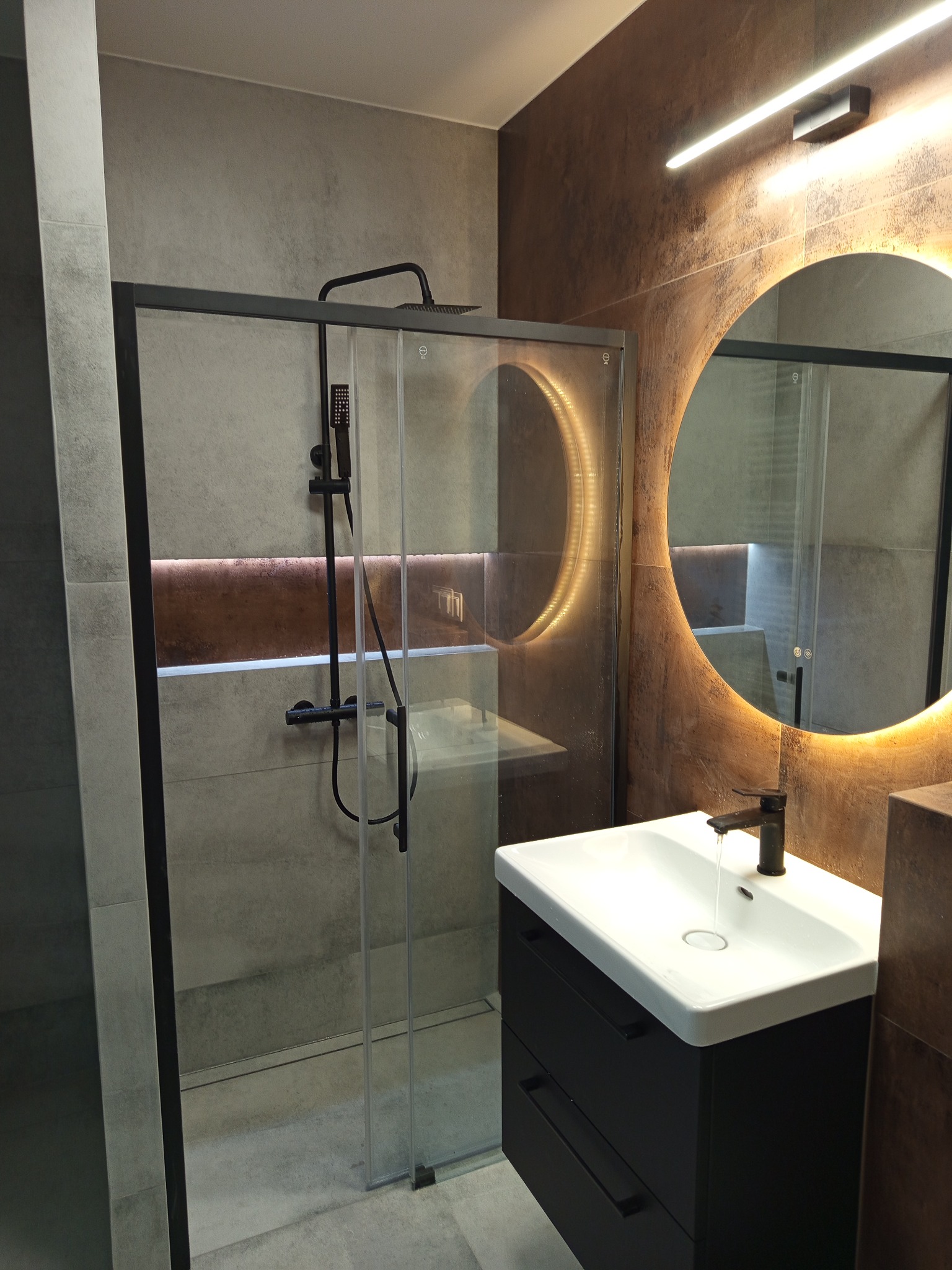 Nowoczesna łazienka z kabiną typu walk-in, czarną armaturą i okrągłym lustrem z podświetleniem LED. Płytki imitujące beton w odcieniach szarości i brązu.