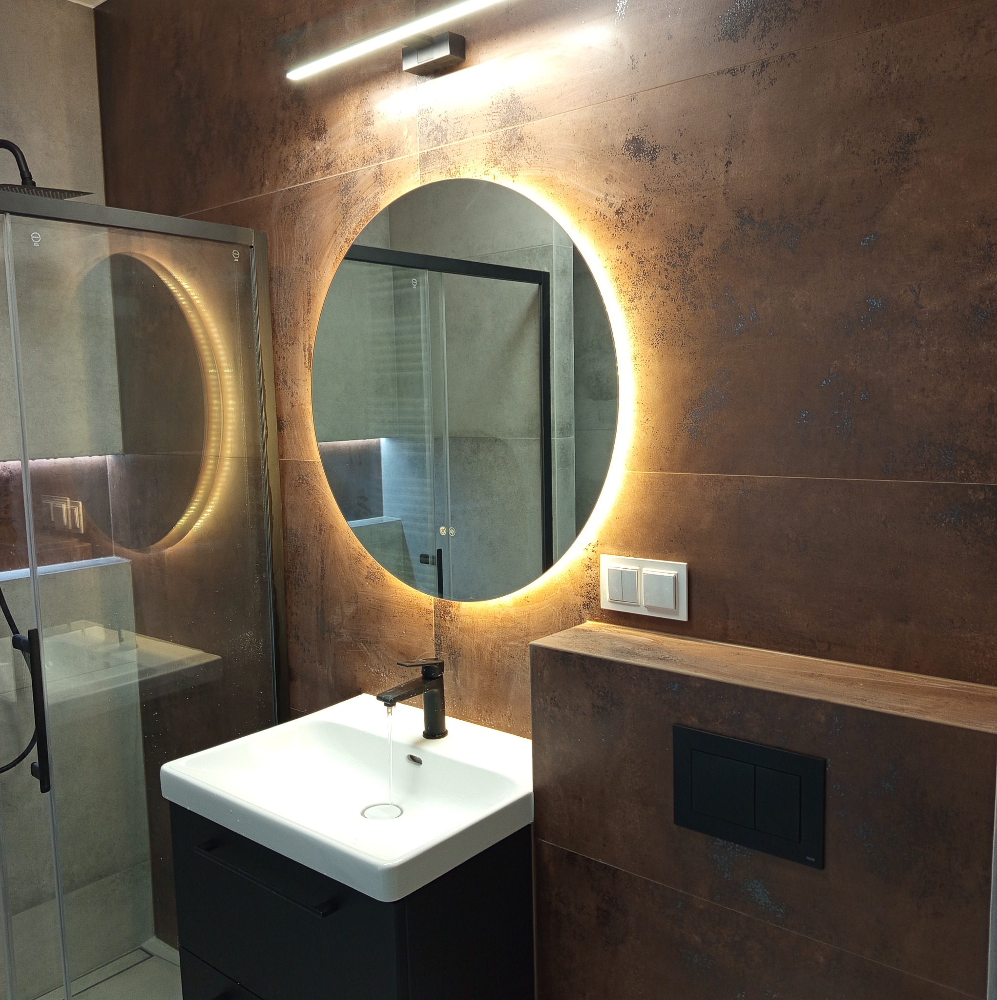 Nowoczesna łazienka z okrągłym lustrem LED, umywalką na szafce i kabiną prysznicową. Brązowe płytki imitujące rdzę na ścianach. Woda leje się z czarnej baterii.