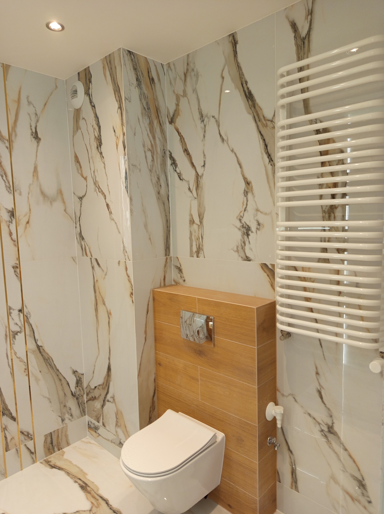 Elegancka łazienka z marmurowymi płytkami, podwieszaną toaletą i grzejnikiem drabinkowym. Beżowe akcenty na tle bieli i złota. Nowoczesny design.