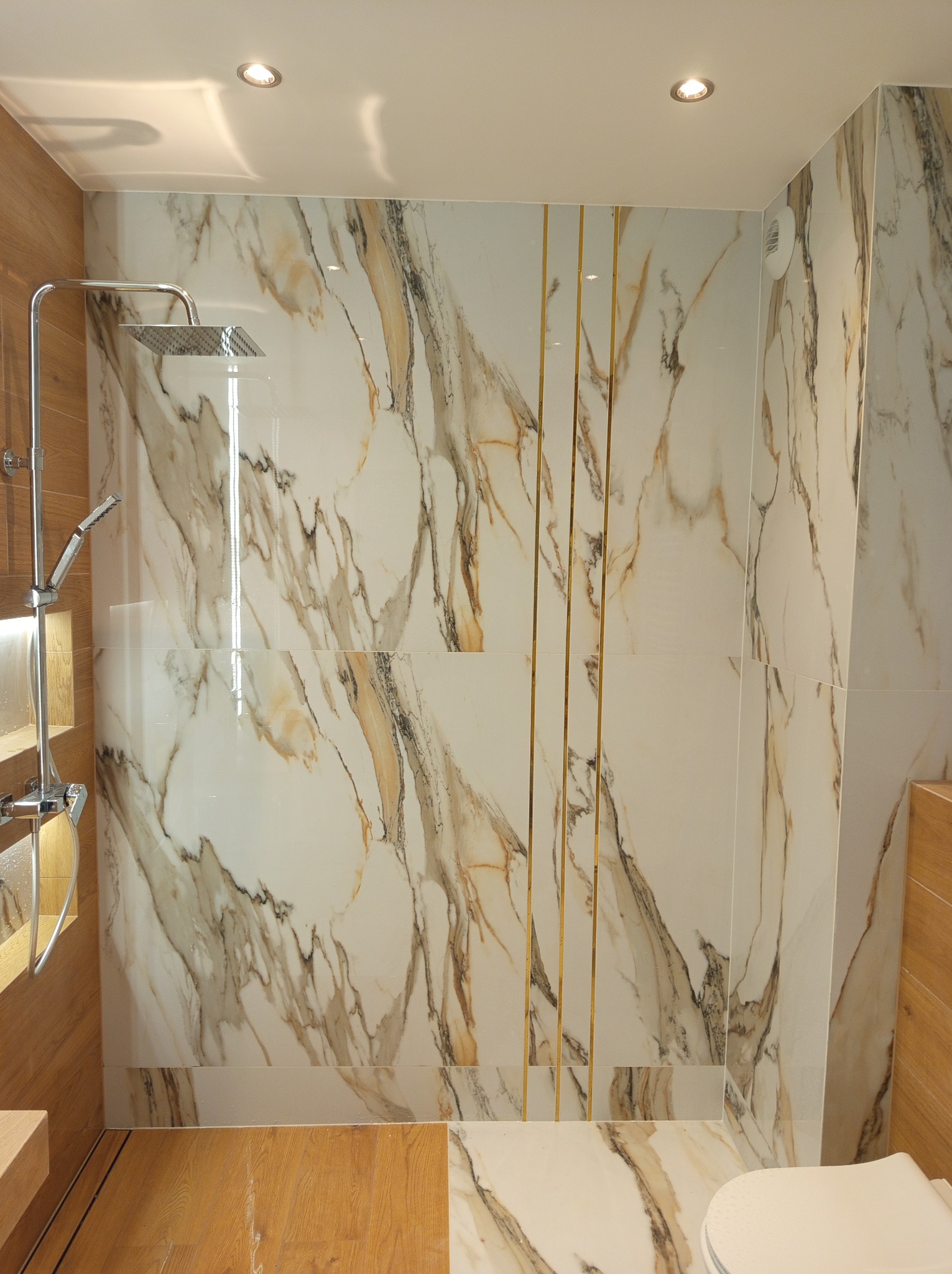 Nowoczesna kabina prysznicowa z marmurowymi płytkami i złotymi akcentami, drewnopodobne wykończenie podłogi i zabudowy. Oświetlenie sufitowe.