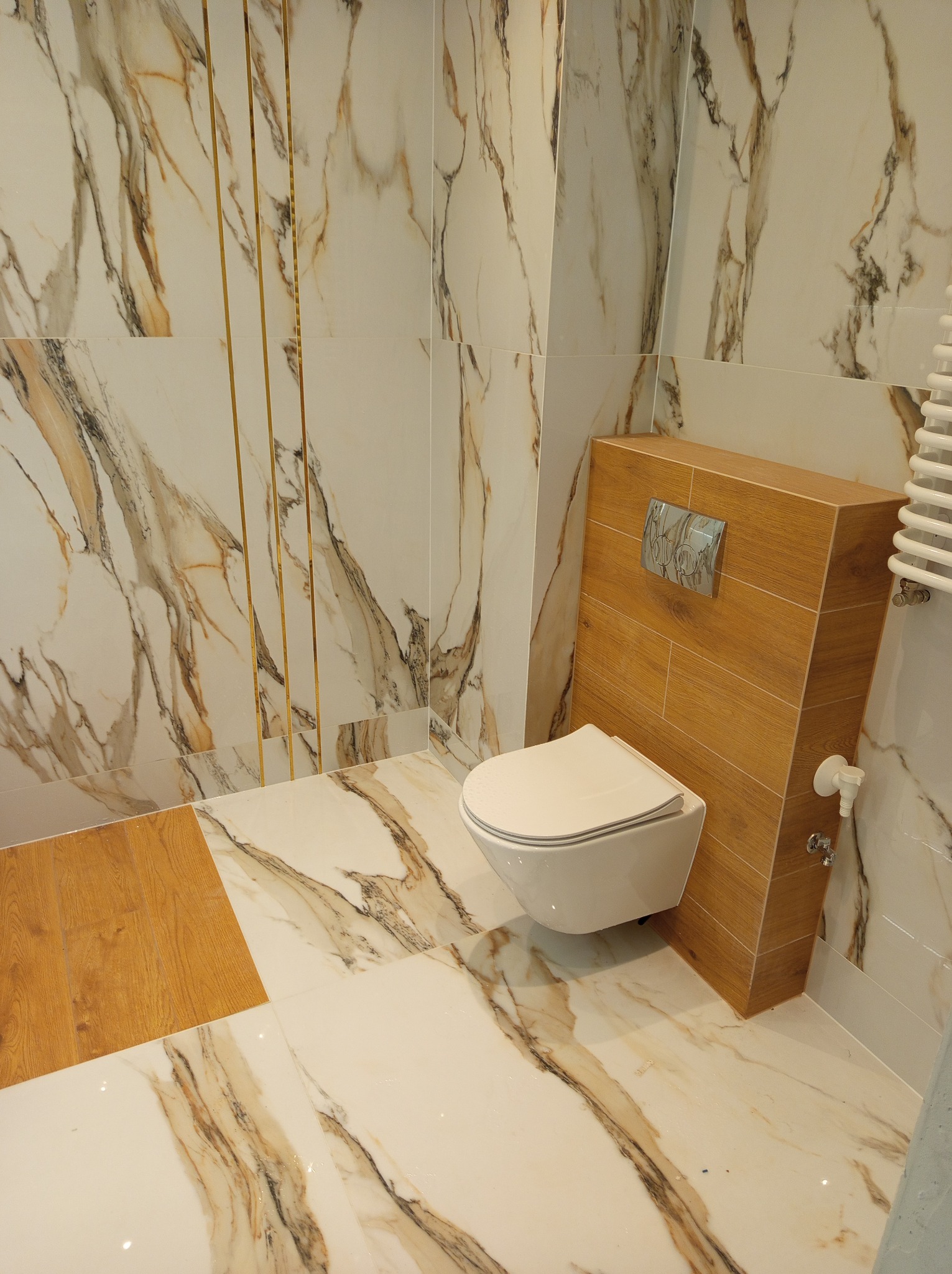 Nowoczesna łazienka z marmurowymi płytkami w odcieniach bieli i brązu, złotymi listwami dekoracyjnymi i podwieszaną toaletą z drewnianą obudową.