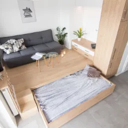 Wielofunkcyjna przestrzeń mieszkalna: szara sofa z poduszkami, szklane stoliki kawowe, wysuwane łóżko z szarą pościelą i drewniane szafki w jasnym, minimalistycznym wnętrzu.