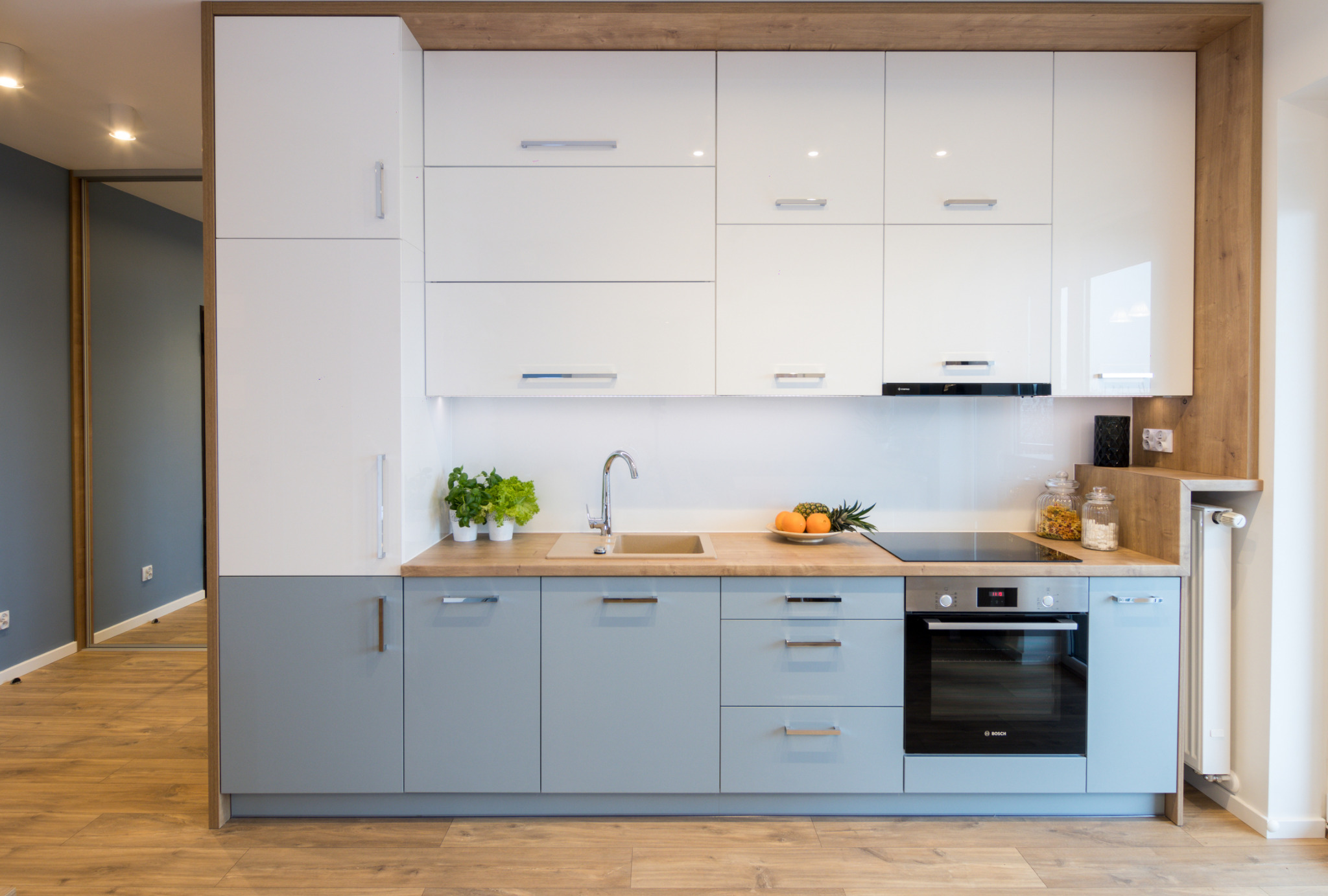 Nowoczesna kuchnia z białymi górnymi szafkami i szarymi dolnymi, drewniany blat z zlewozmywakiem i owocami, piekarnik Bosch w zabudowie, podłoga drewnopodobna, widoczne rośliny w doniczkach.