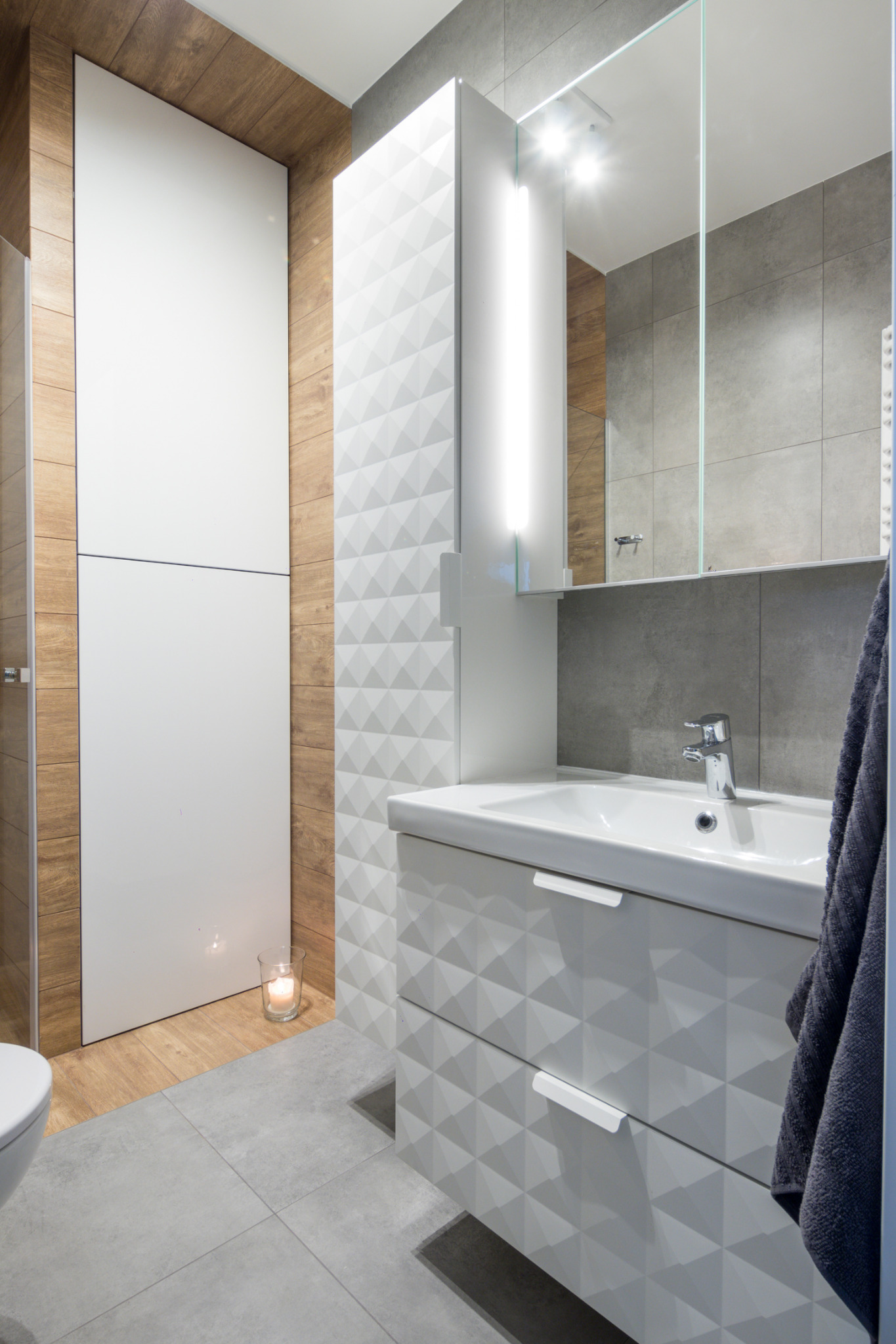 Nowoczesna łazienka z białą szafką umywalkową o geometrycznym wzorze 3D, podświetlanym lustrem, drewnopodobnymi panelami ściennymi i zapaloną świecą na podłodze.