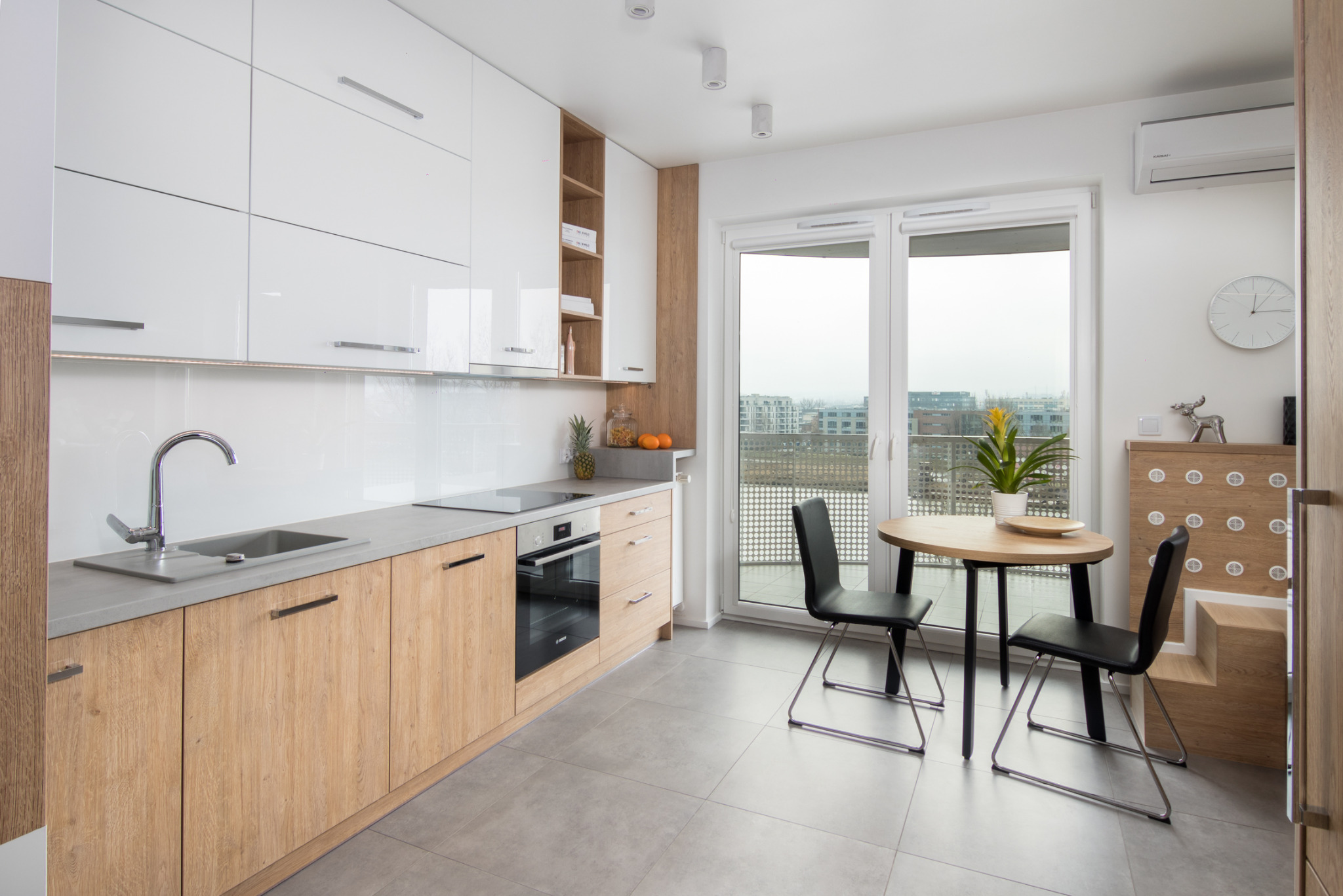 Jasna kuchnia z białymi lakierowanymi szafkami górnymi i drewnianymi dolnymi, szary blat, okrągły stół z czarnymi krzesłami, widok na miasto z balkonu.