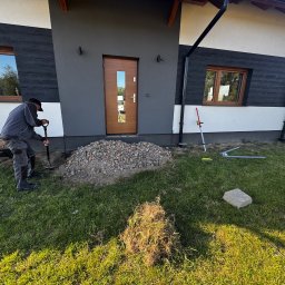 Hubert Mańczak Usługi Brukarskie - Pracownik z łopatą przygotowuje podłoże pod kostkę obok nowoczesnego domu z drewnianymi oknami i drzwiami. Widoczny pryzmat gruzu i trawa.