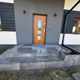 Hubert Mańczak Usługi Brukarskie - Schody wejściowe wyłożone szarą kostką brukową prowadzące do nowoczesnego domu z drewnianymi oknami i drzwiami. Widoczny fragment elewacji i trawnik.