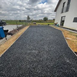 Ukończona powierzchnia z czarnego kruszywa wypełniającego plastikowe kratki, obrzeża betonowe. Dom z białym tynkiem w tle, pochmurne niebo nad posesją.