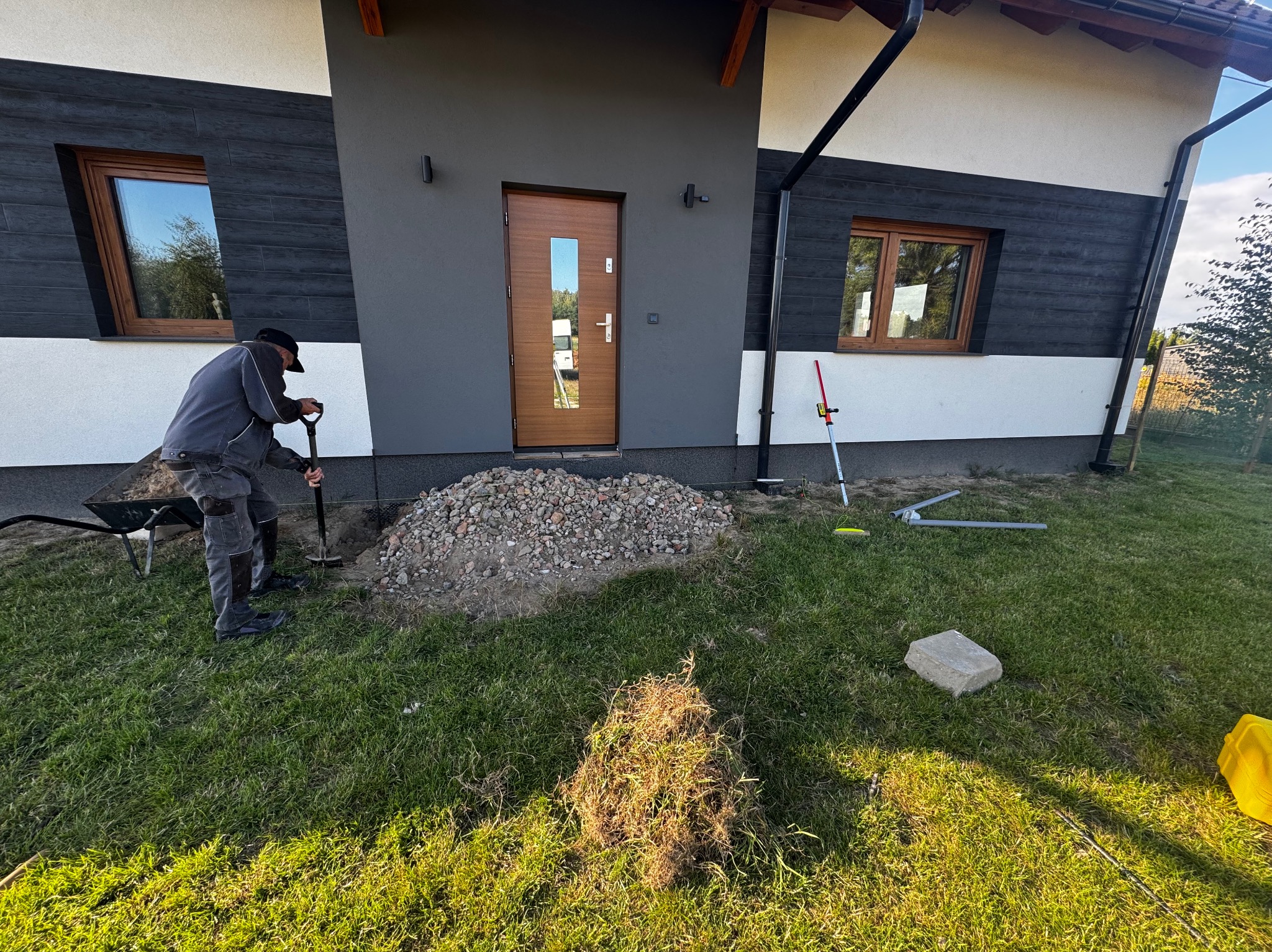 Pracownik z łopatą przygotowuje podłoże pod kostkę obok nowoczesnego domu z drewnianymi oknami i drzwiami. Widoczny pryzmat gruzu i trawa.