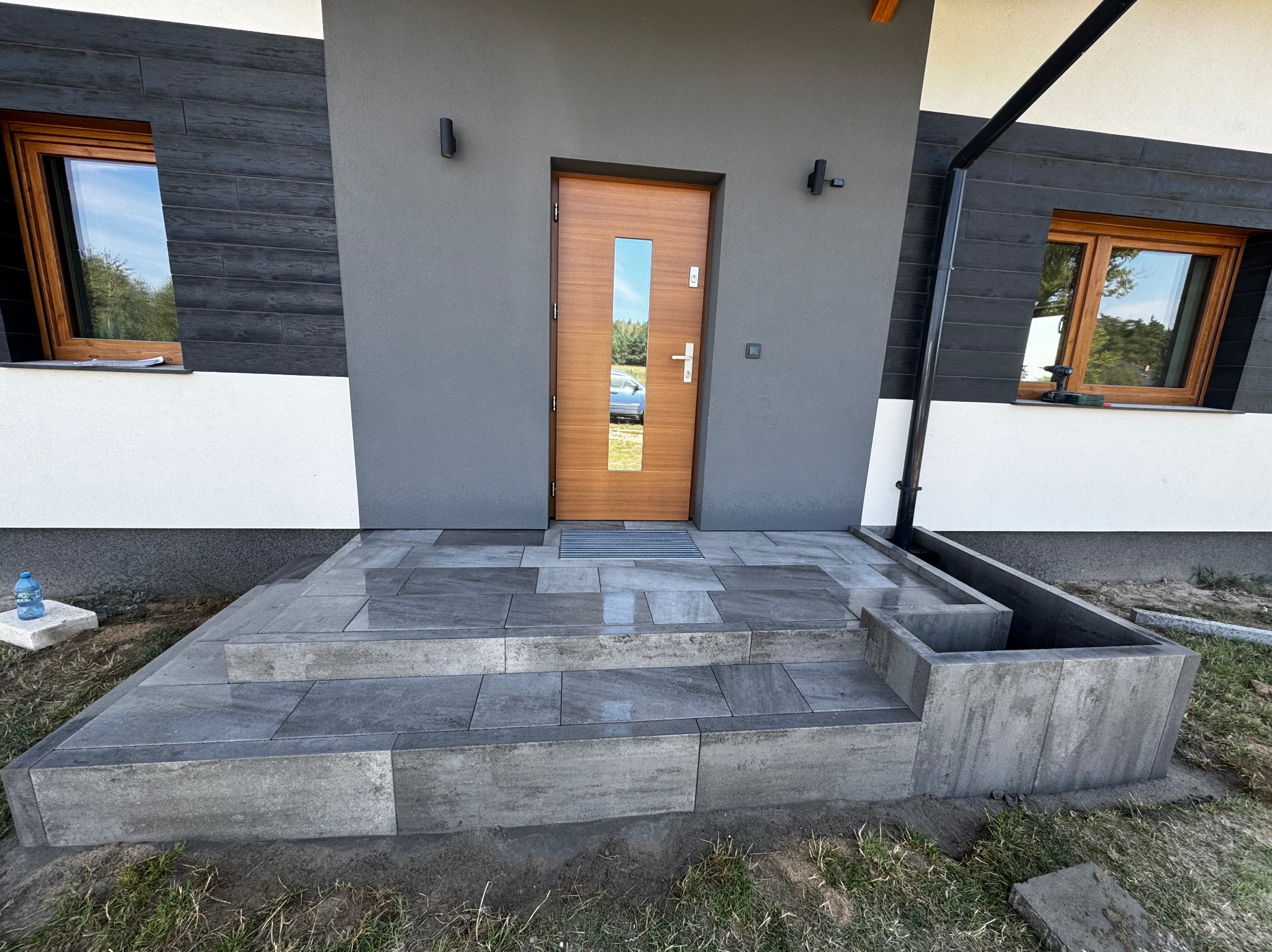 Schody wejściowe wyłożone szarą kostką brukową prowadzące do nowoczesnego domu z drewnianymi oknami i drzwiami. Widoczny fragment elewacji i trawnik.